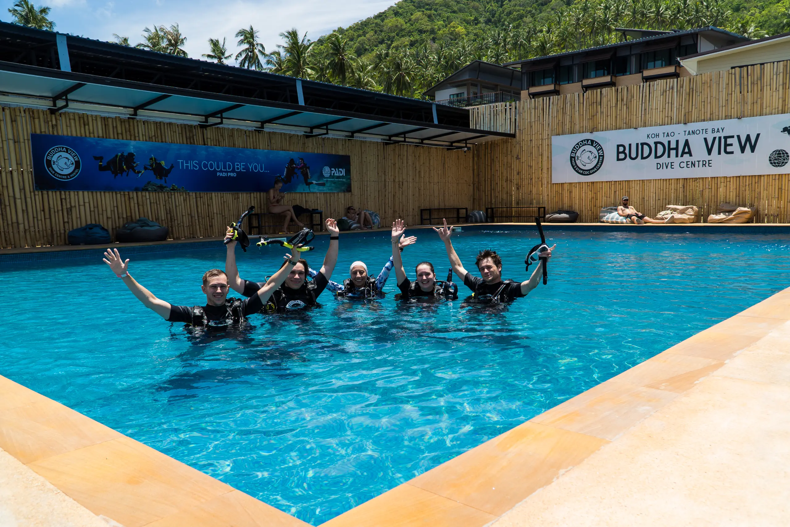Cinco alumnos en neopreno de pie en la piscina de entrenamiento de Buddha View, con los brazos en alto y sus aletas y tubos, y el cartel del centro de buceo detrás